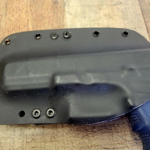 Peut inclure: Un étui en polymère noir moulé avec plusieurs trous de vis. L'étui est conçu pour s'adapter à une arme de poing, avec une poignée en polymère bleu visible. L'étui est présenté sur une surface en bois.