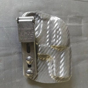 K&ouml;nnte beinhalten: Ein schwarzes und graues Carbonfaser-Holster mit einem schwarzen Metallclip. Der Clip tr&auml;gt den Text "Belt Clip".