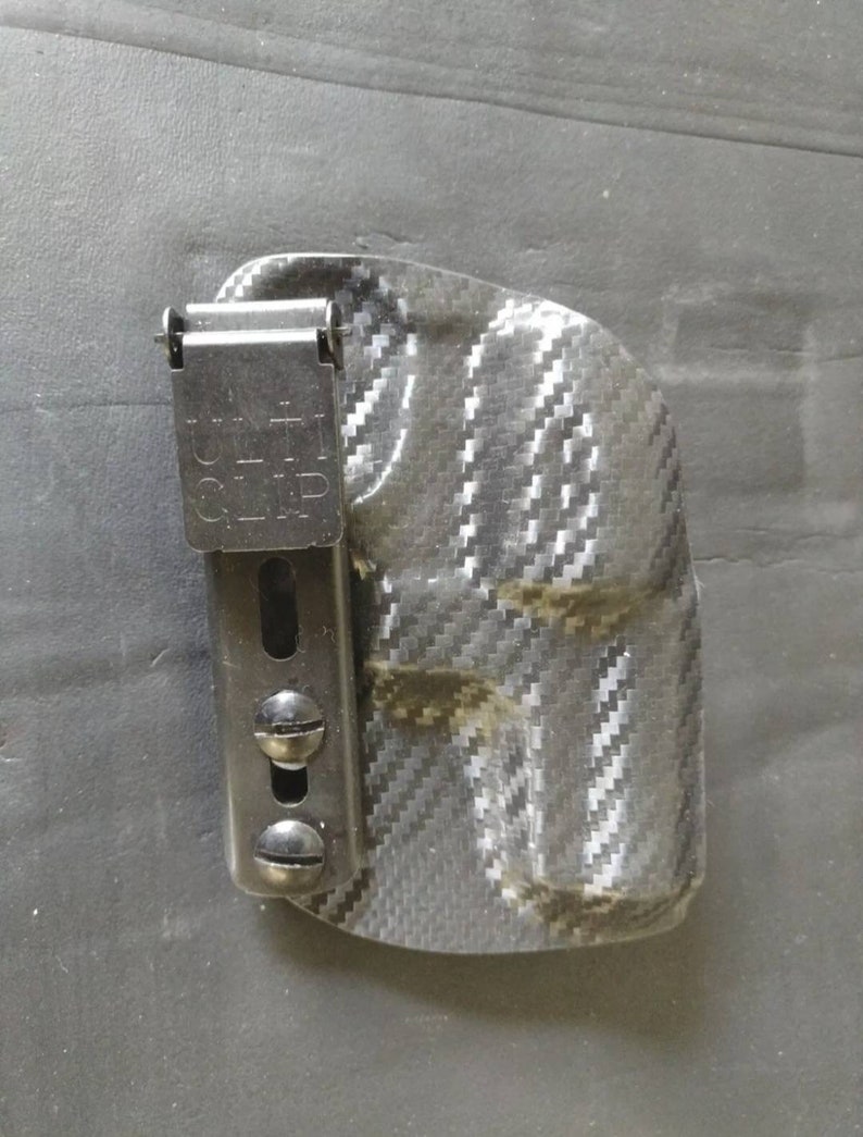 K&ouml;nnte beinhalten: Ein schwarzes und graues Carbonfaser-Holster mit einem schwarzen Metallclip. Der Clip tr&auml;gt den Text "Belt Clip".