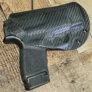 May include: A black carbon fiber holster with a textured grip. The holster is designed to securely hold a handgun. The holster has a carbon fiber weave pattern and is mounted on a wooden surface.