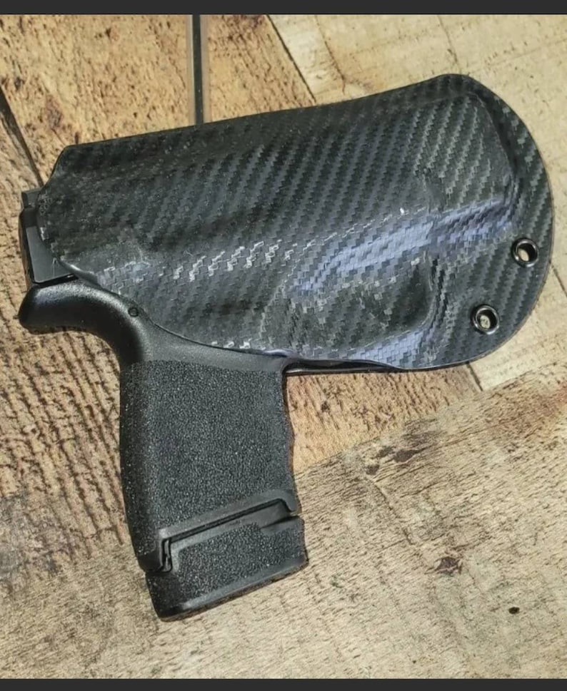 May include: A black carbon fiber holster with a textured grip. The holster is designed to securely hold a handgun. The holster has a carbon fiber weave pattern and is mounted on a wooden surface.