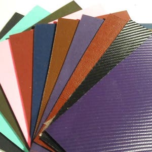May include: A fan of various colored leather and textured vinyl samples. Colors include black, teal, pink, red, brown, navy, purple, and carbon fiber. The materials are arranged in a semi-circular pattern, showcasing their diverse textures and hues.
