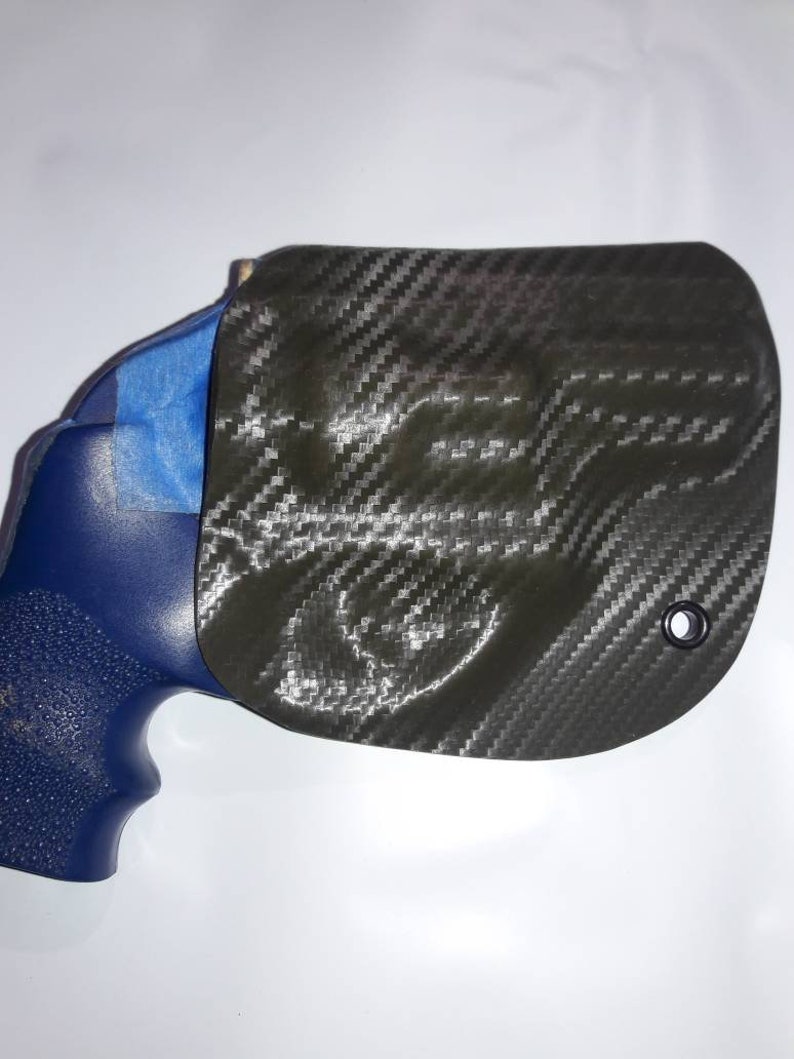 May include: A black carbon fiber holster with a blue gun inside. The holster has a small hole on the top right side.