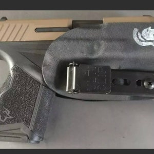 May include: A black handgun with a tan slide is secured in a black holster. The holster features a silver clip and a white graphic of a wolf. The grip of the gun has a textured surface.