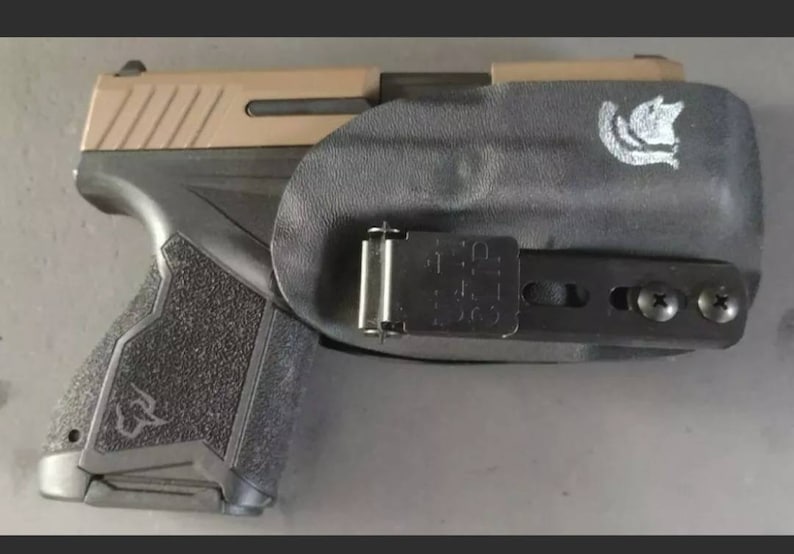 May include: A black and tan handgun with a black holster. The holster has a black clip and two screws on the side. The holster has a logo on the top.