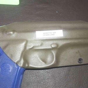 May include: A green leather holster with a label that reads "Beretta 92F Rowb-LWB". The holster has two black eyelets on the side. A blue and black handgun is partially visible in the image.