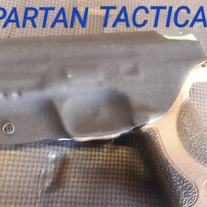May include: Black holster with the text "SPARTAN TACTICAL" in blue. The holster is covering a tan handgun. The image is taken on a textured surface.