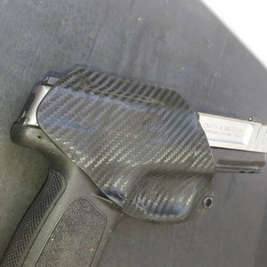 May include: A black carbon fiber holster with a silver handgun inside. The handgun has the text "SMITH & WESSON SPRINGFIELD, MA, USA" on the side.