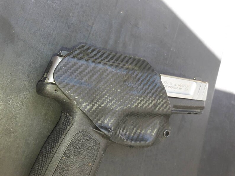 May include: A black carbon fiber holster with a silver handgun inside. The handgun has the text "SMITH & WESSON SPRINGFIELD, MA, USA" on the side.