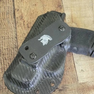 May include: Black carbon fiber holster with a Spartan helmet emblem. The holster is designed to hold a handgun. The holster has a textured surface and is secured with screws. The background is a wooden surface.