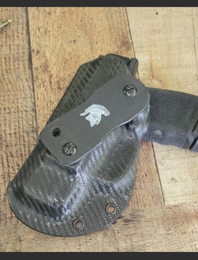 May include: Black carbon fiber holster with a Spartan helmet emblem. The holster is designed to hold a handgun. The holster has a textured surface and is secured with screws. The background is a wooden surface.