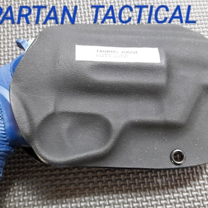 May include: Black leather holster with a blue Taurus Judge pistol inside. The holster has two black snaps on the side and the text "Taurus Judge" is printed on a white label.