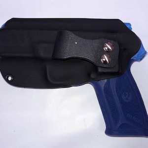 Peut inclure: Un &eacute;tui en cuir noir avec une boucle de ceinture rivet&eacute;e argent&eacute;e. L'&eacute;tui recouvre une arme de poing bleue. Le mot "Ruger" est visible sur la poign&eacute;e. L'&eacute;tui a un seul &oelig;illet en bas &agrave; gauche.