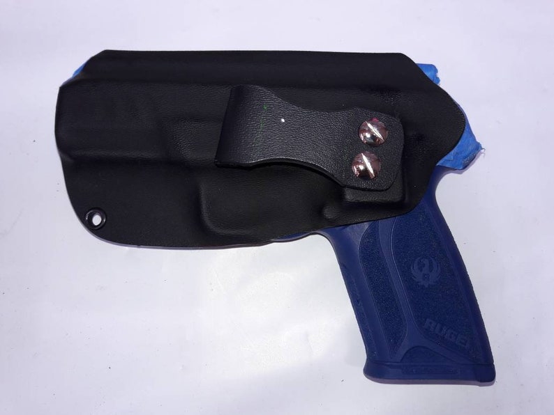Peut inclure: Un &eacute;tui en cuir noir avec une boucle de ceinture rivet&eacute;e argent&eacute;e. L'&eacute;tui recouvre une arme de poing bleue. Le mot "Ruger" est visible sur la poign&eacute;e. L'&eacute;tui a un seul &oelig;illet en bas &agrave; gauche.