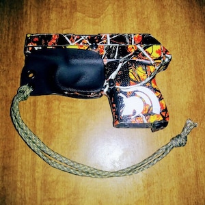 Puede incluir: Una pistola con estampado de camuflaje negro y naranja con una funda negra y un cordón verde y amarillo.