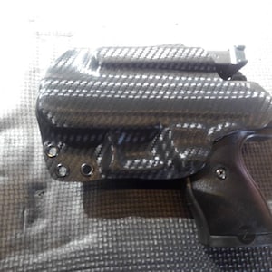 May include: A black and gray gun holster with a carbon fiber pattern. The holster is designed to fit a handgun with a black polymer grip.