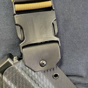 May include: Close-up of a black holster with a carbon fibre pattern, featuring a black buckle and tan strap. The holster has several eyelets and is attached to a black buckle.