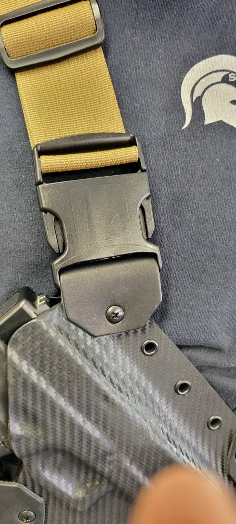 May include: Close-up of a black holster with a carbon fibre pattern, featuring a black buckle and tan strap. The holster has several eyelets and is attached to a black buckle.