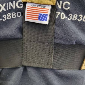 May include: Close-up of a dark grey fabric with a tan strap and a label featuring the American flag. The label reads "Made in the USA". The strap is stitched with a black thread.