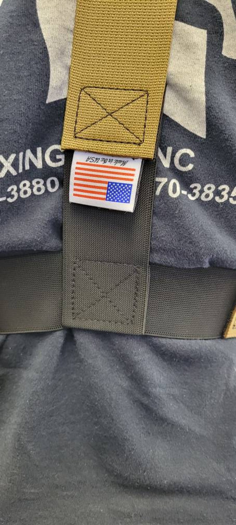 May include: Close-up of a dark grey fabric with a tan strap and a label featuring the American flag. The label reads "Made in the USA". The strap is stitched with a black thread.