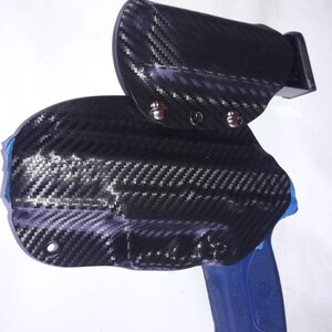 Puede incluir: Una funda para pistola de fibra de carbono negra y gris con un mango de pistola azul. La funda tiene un cierre a presión y está diseñada para llevar oculta.