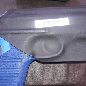 K&ouml;nnte beinhalten: Ein schwarzes Lederholster mit einem blauen Klebebandrand. Das Holster tr&auml;gt ein Etikett mit der Aufschrift "RUGER P95/PRS  BOTW L1TW".
