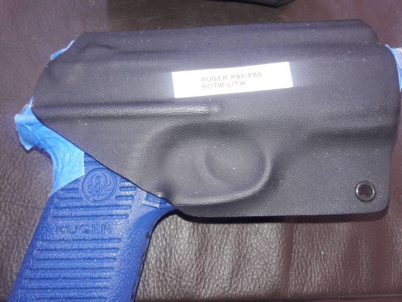 K&ouml;nnte beinhalten: Ein schwarzes Lederholster mit einem blauen Klebebandrand. Das Holster tr&auml;gt ein Etikett mit der Aufschrift "RUGER P95/PRS  BOTW L1TW".