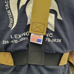 May include: Tan and black tactical belt system. The belt features a tan shoulder strap and a black waist belt. The label with the American flag is visible. The shirt in the background has the text "SPARTAN TACTICAL HOLSTERS & HS".