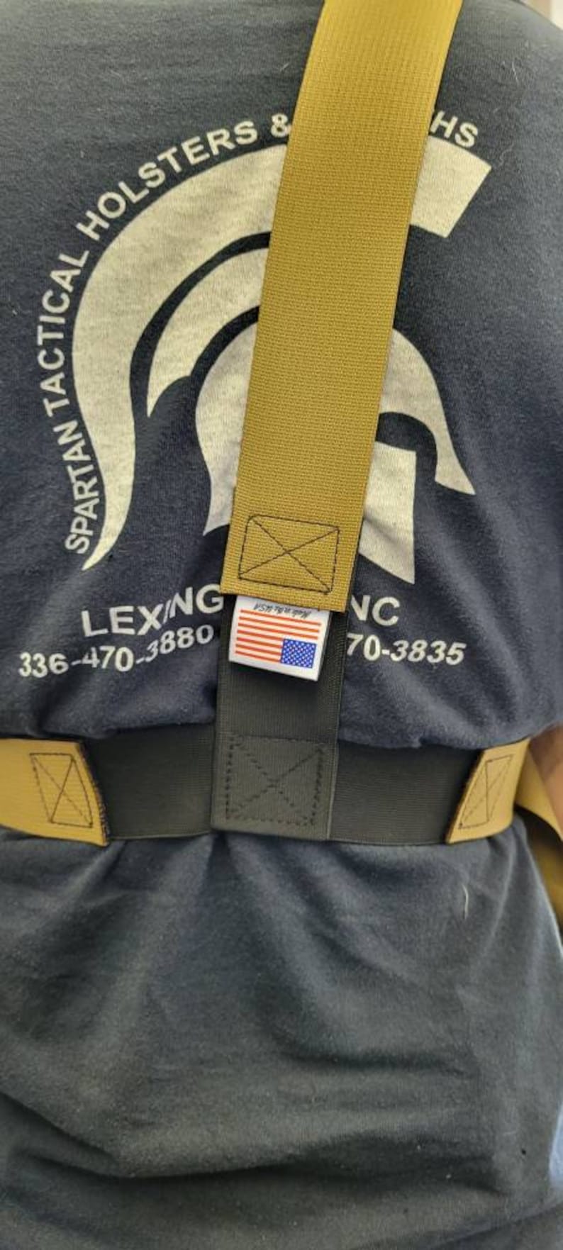 May include: Tan and black tactical belt system. The belt features a tan shoulder strap and a black waist belt. The label with the American flag is visible. The shirt in the background has the text "SPARTAN TACTICAL HOLSTERS & HS".