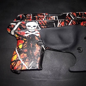 May include: A compact handgun with a skull and crossbones design on the grip. The gun features a black holster. The frame has a red, orange, and black camouflage pattern.