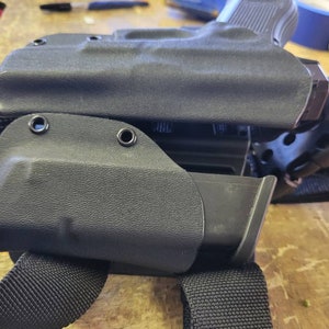 May include: A black leather holster with a magazine pouch attached to a black nylon strap. The holster has two metal eyelets on the side.