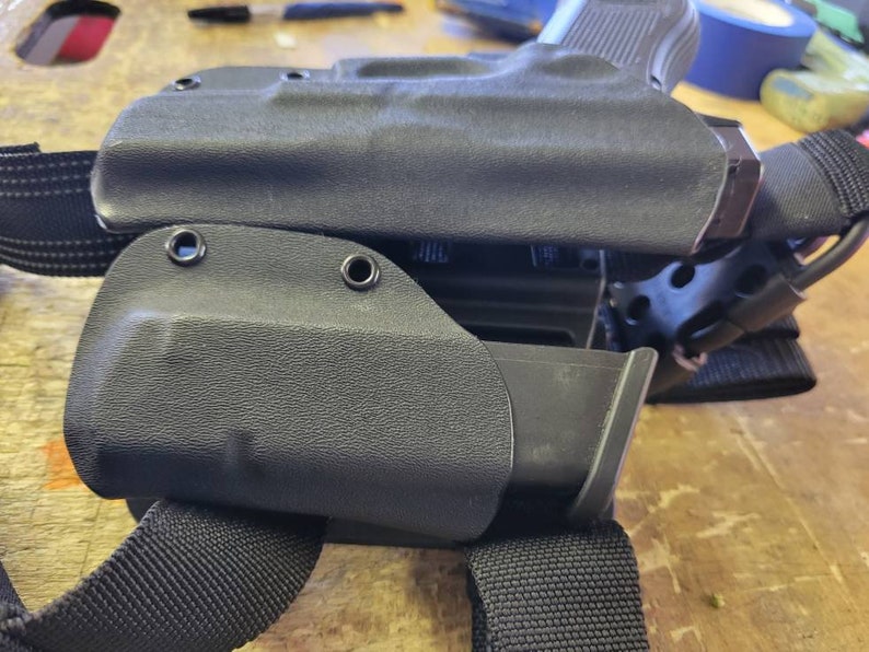 May include: A black leather holster with a magazine pouch attached to a black nylon strap. The holster has two metal eyelets on the side.