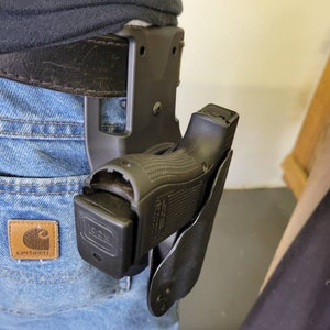 May include: A black Glock handgun in a black holster attached to a brown leather belt. The holster is secured to the belt, and the gun is partially visible. The jeans have a Carhartt patch.