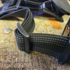 May include: Close-up of a black adjustable strap with a textured fabric and a plastic buckle.