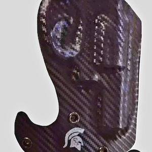 May include: A black and grey carbon fiber holster with a Spartan helmet logo. The holster has a hook on the bottom and multiple small holes around the edges.