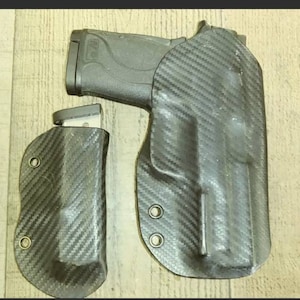 May include: A black handgun with a magazine and two carbon fiber holsters. The holsters are designed to hold the handgun and magazine securely. The carbon fiber material has a woven pattern.