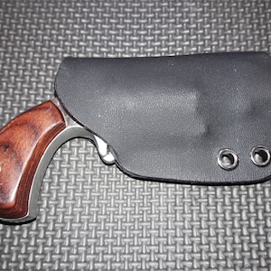 Peut inclure: Un petit pistolet argenté avec une poignée en bois est dans un étui en cuir noir. L'étui a deux œillets métalliques.