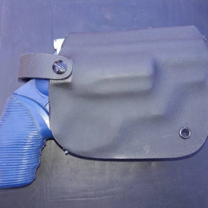 Op de afbeelding: Een blauw en zwart pistool met een zwart leren holster. Het holster heeft een band en een metalen oog.