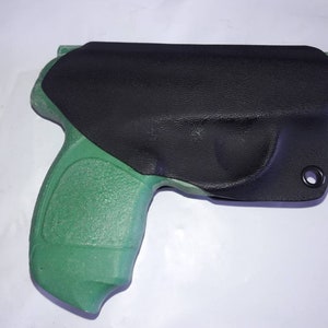 May include: A black holster with a textured surface covers a green pistol grip. The holster has a single eyelet and is designed to fit the shape of the firearm. The image is taken on a white background.