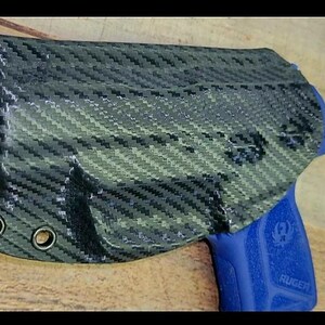 K&ouml;nnte beinhalten: Eine schwarze und graue Kohlefaserholster mit drei &Ouml;sen. Eine blaue Handfeuerwaffe ist teilweise in der Holster sichtbar. Die Holster liegt auf einer Holzoberfl&auml;che.