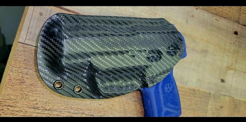 K&ouml;nnte beinhalten: Eine schwarze und graue Kohlefaserholster mit drei &Ouml;sen. Eine blaue Handfeuerwaffe ist teilweise in der Holster sichtbar. Die Holster liegt auf einer Holzoberfl&auml;che.