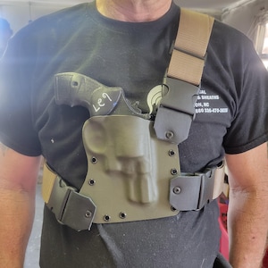 May include: A chest holster system in olive green with a tan strap and belt. The holster is designed to hold a revolver. The system has black buckles and is worn over a black t-shirt.