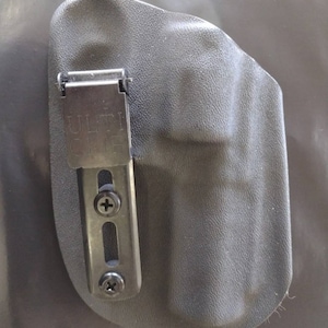 May include: A black holster with a textured surface and a metal clip. The clip is marked "ULTI CLIP" and is attached with screws. The holster is designed to hold a firearm and is made of durable material.