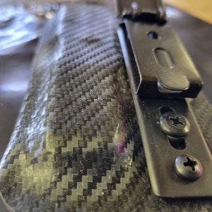 May include: Close-up of a black carbon fiber holster with a metal clip and screws. The holster has a textured, woven pattern. The metal clip is attached with two screws.