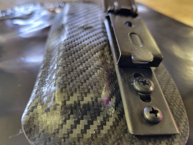 May include: Close-up of a black carbon fiber holster with a metal clip and screws. The holster has a textured, woven pattern. The metal clip is attached with two screws.