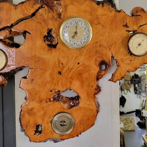 May include: A wooden wall clock with a natural wood grain finish. The clock has three round faces, one with a clock face, one with a barometer, and one with a thermometer. The clock is mounted on a piece of wood that looks like a tree slice.