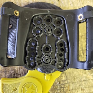 Puede incluir: Un holster de pl&aacute;stico negro con puntos de montaje ajustables para una peque&ntilde;a pistola amarilla con una estrella en el lateral.