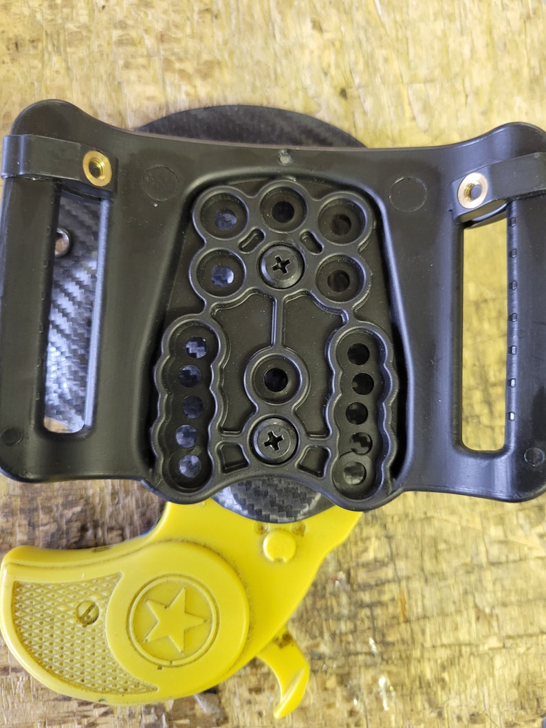 Puede incluir: Un holster de pl&aacute;stico negro con puntos de montaje ajustables para una peque&ntilde;a pistola amarilla con una estrella en el lateral.
