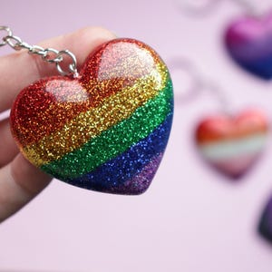 Könnte beinhalten: Ein herzförmiger Schlüsselanhänger mit einem Regenbogen-Glitzer-Design, gehalten vor einem verschwommenen Hintergrund anderer herzförmiger Schlüsselanhänger. Das Regenbogen-Design weist rote, orange, gelbe, grüne, blaue und violette Glitzer auf. Der Schlüsselanhänger ist an einer silbernen Kette befestigt.