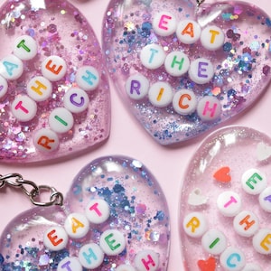 Könnte beinhalten: Vier herzförmige Schlüsselanhänger mit dem Schriftzug "EAT THE RICH" aus bunten Buchstabenperlen. Die transparenten Harzherzen sind mit Glitzer und kleinen dekorativen Elementen gefüllt und an silbernen Schlüsselanhängern befestigt.